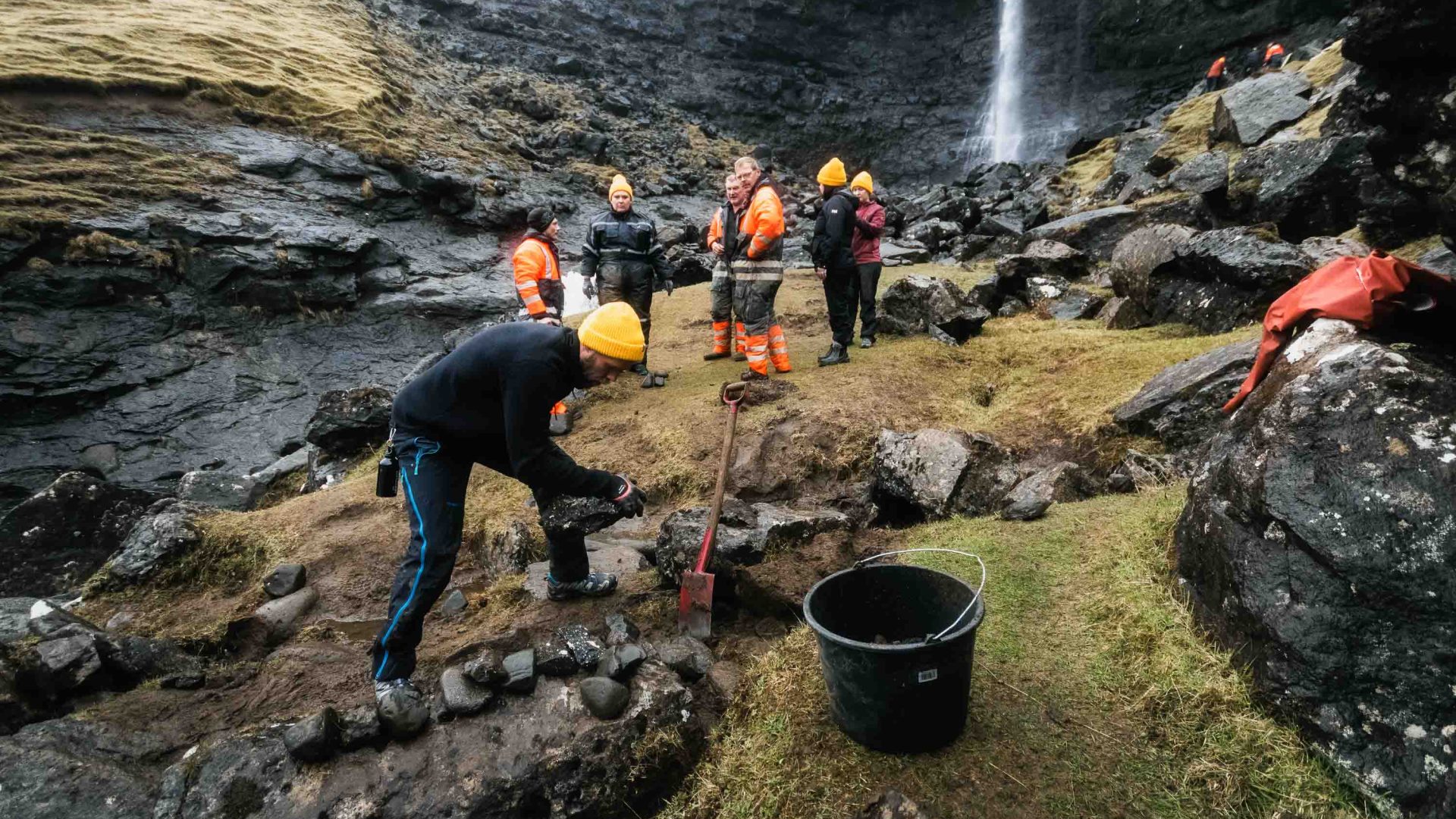 A volunteer digs into the ground near a waterfall while a group of others look on.